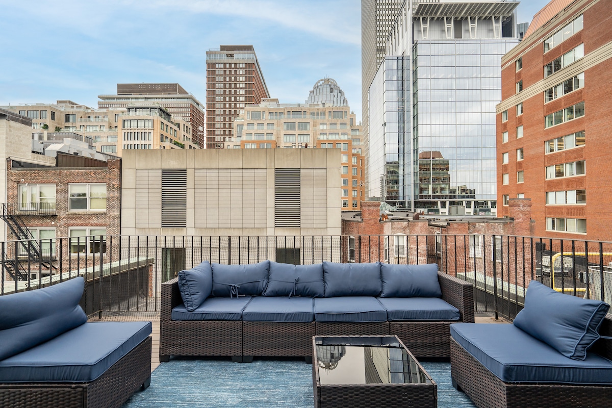A private rooftop deck features spacious outdoor seating with plush navy cushions arranged around a glass coffee table. The backdrop showcases a cityscape filled with modern buildings and classic architecture, creating a vibrant atmosphere for relaxation.
