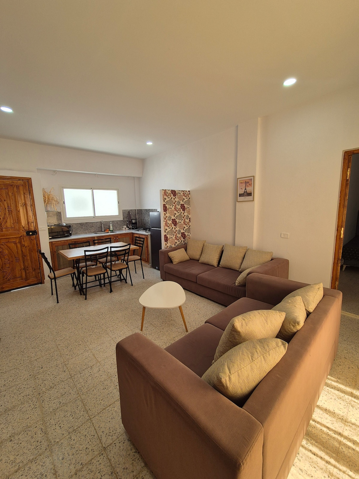 The living area features two sofas facing each other, accompanied by a small coffee table. A dining table with multiple chairs is positioned nearby, and natural light enters through a window above the kitchen area, enhancing the bright and spacious atmosphere.
