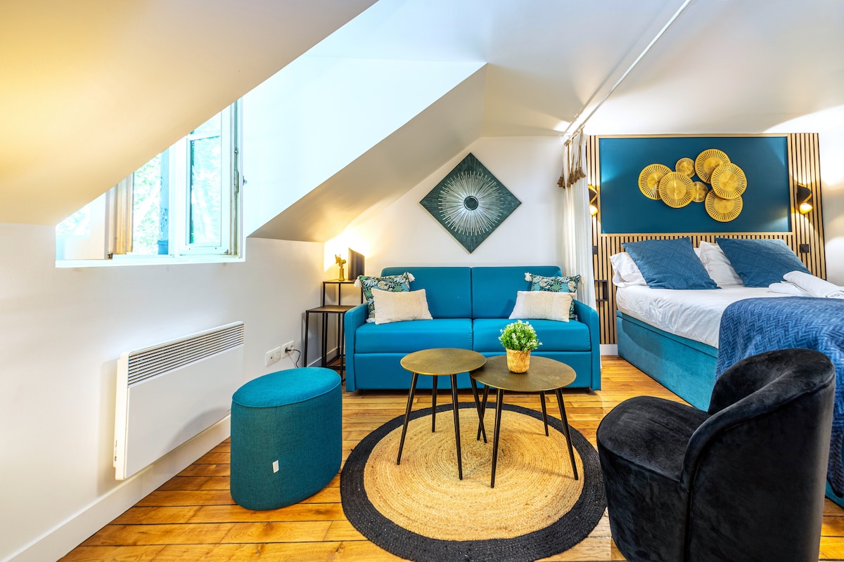 A spacious studio features a comfortable sleeping area with a queen-size bed and a convertible sofa. Two round tables are placed on a textured rug, complemented by stylish chairs. Natural light enters through windows, illuminating the modern decor with blue accents and artistic wall art.