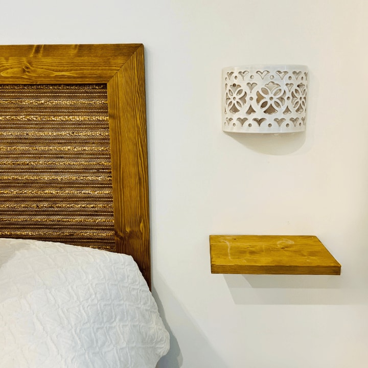 A wooden headboard featuring textured details is positioned above a cozy bed. A small wooden shelf is attached to the wall alongside an intricately designed white wall lamp, providing decorative elements and functionality in the sleeping area.