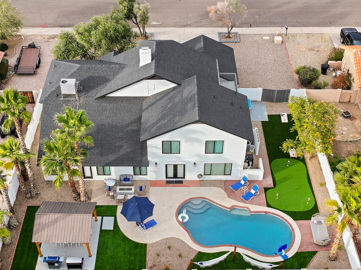 An aerial view of a contemporary home reveals a spacious layout with a heated swimming pool and surrounding lounge chairs. A green putting green and a shaded area with a cabana are visible in the backyard, complemented by palm trees adding greenery to the landscape.