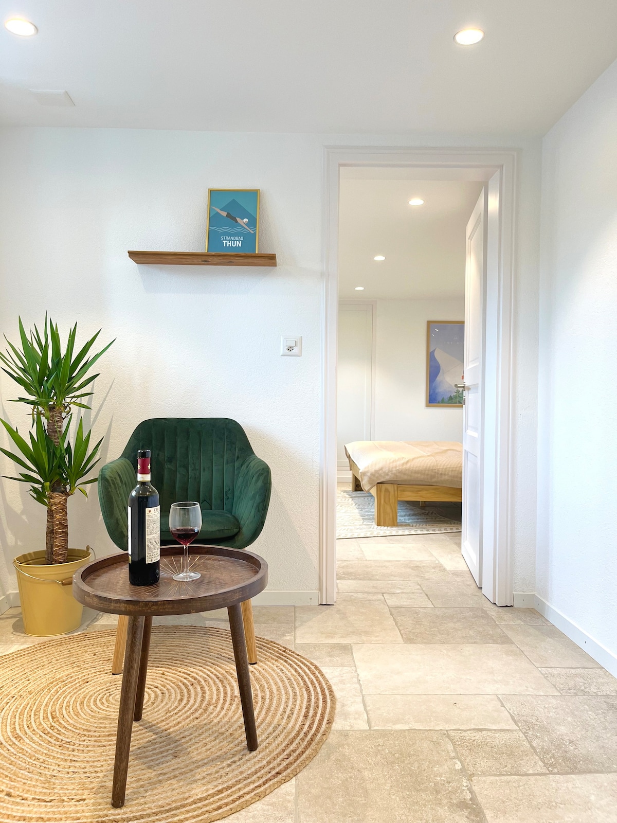 A cozy corner features a green upholstered chair next to a small round table with a bottle of wine and a glass. A potted plant adds a touch of greenery, while a doorway leads to a bright sleeping area in the background.
