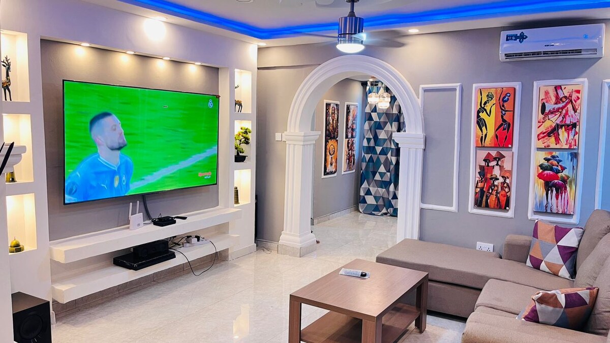 A modern living room features a large wall-mounted flat-screen TV, flanked by decorative art pieces. Comfortable seating arrangements include a plush sofa and a wooden coffee table. Ambient blue lighting highlights the ceiling and enhances the overall design, creating a warm and inviting space.