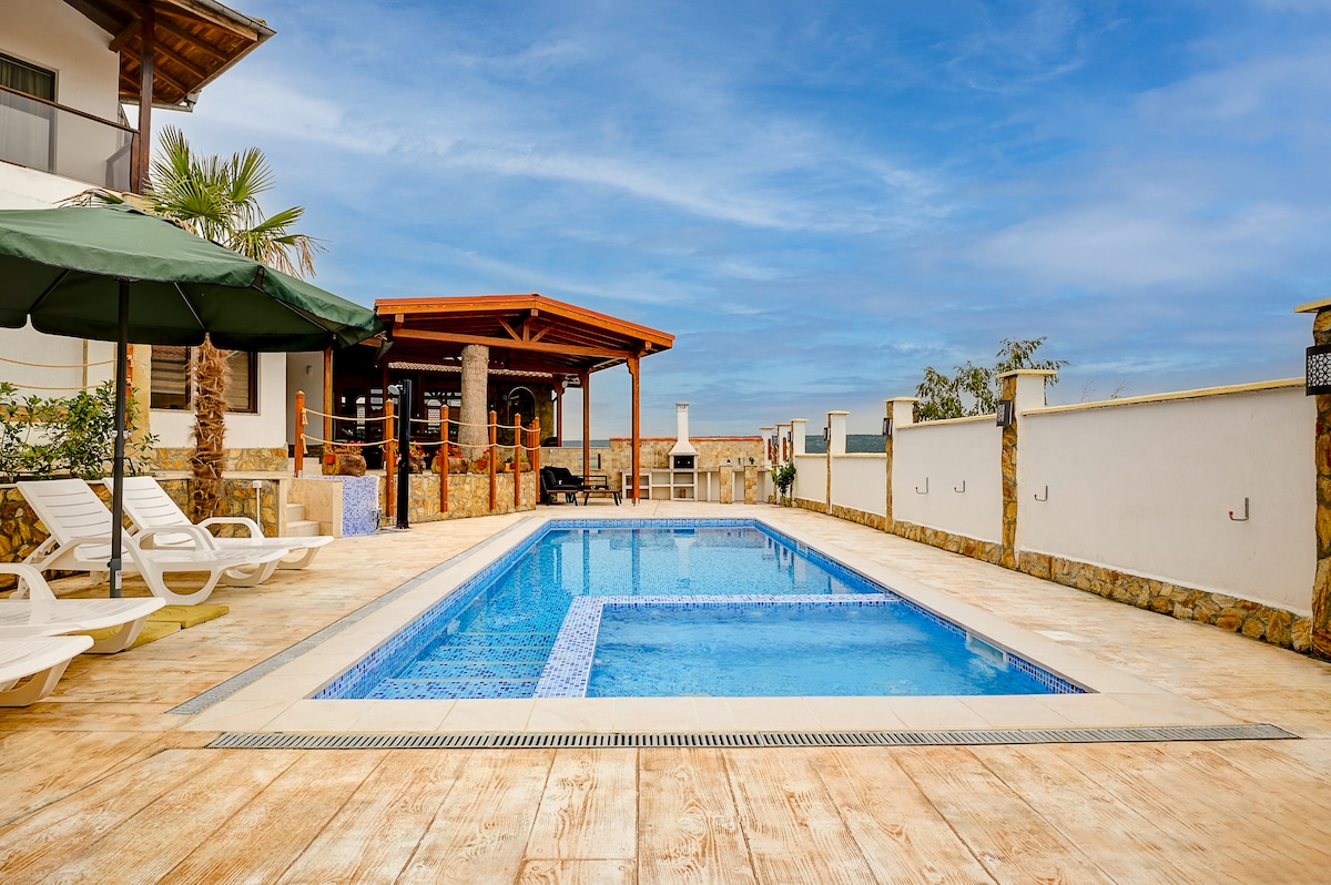 A spacious outdoor area features a heated swimming pool with a jacuzzi section. Sun loungers are positioned beside the pool, accompanied by a shaded seating area under an umbrella. A covered terrace with wooden beams overlooks the pool, enhancing the serene environment.