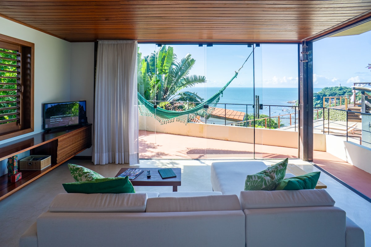 A spacious living area includes a modern sofa adorned with cushions in varying shades of green. Large glass doors open onto a terrace with ocean views, where a hammock hangs invitingly. Lush greenery is visible in the background, enhancing the natural setting.