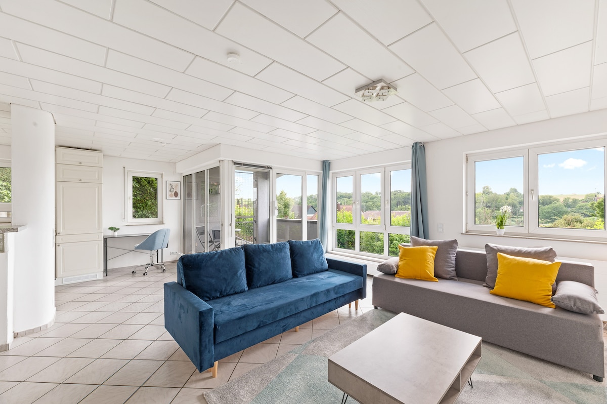 A spacious living area is filled with natural light, featuring two large sofas with yellow accent cushions. A coffee table sits centrally, with a viewing area alongside large windows offering green landscape views. A desk is positioned nearby for work needs.