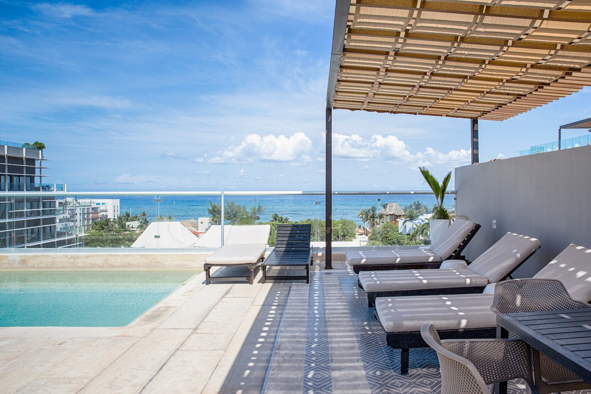A rooftop area features a tranquil pool and several lounge chairs arranged under a shaded pergola. A coastal view is visible in the background, with blue skies and distant palm trees enhancing the serene atmosphere.