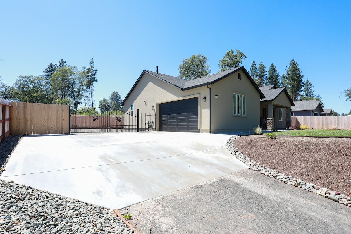 A welcoming exterior view of a modern home highlights a spacious driveway and well-maintained landscaping. The structure features light-colored walls and a black garage door. Surrounding trees provide a natural backdrop, while a fenced yard enhances privacy and security.