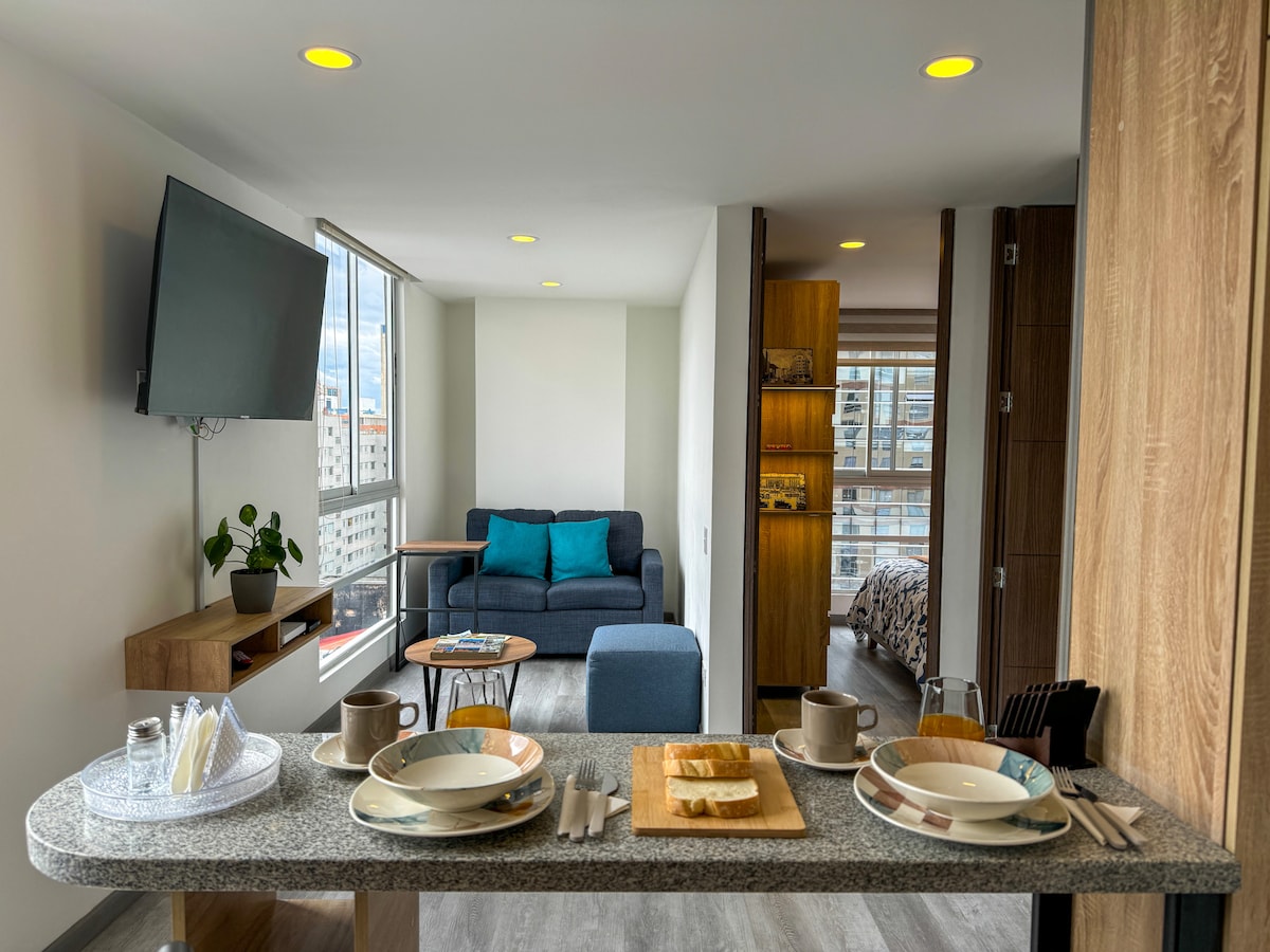 The image showcases a modern open-concept living space featuring a dining area with a set of plates and cups arranged on a countertop. A comfortable sofa with teal cushions can be seen in the background, along with a well-lit bedroom space visible through a partition.
