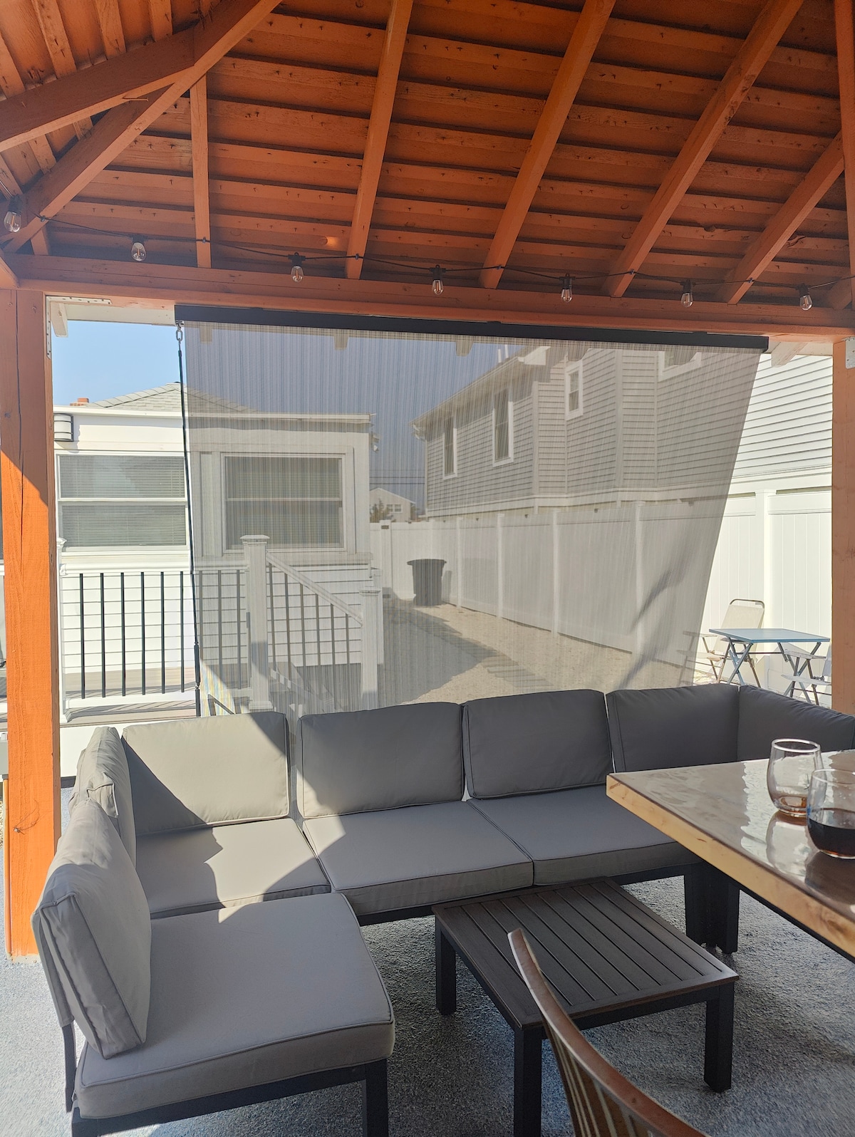 A shaded outdoor pavilion features a comfortable sectional sofa in neutral tones arranged around a wooden table. Sheer privacy screens flank the space, and sunlight filters through the roof’s beams, creating a pleasant ambiance. The setting invites relaxation and social gatherings.