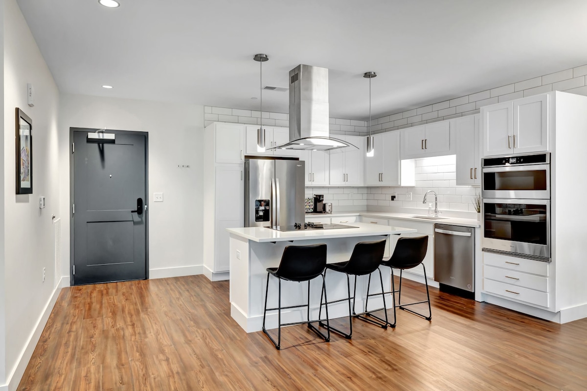 A modern kitchen features stainless steel appliances, including a refrigerator and double ovens. A spacious island with seating for four is complemented by sleek cabinetry and white countertops. Warm wood flooring adds a cozy touch, while a doorway leads to the entrance of the unit.