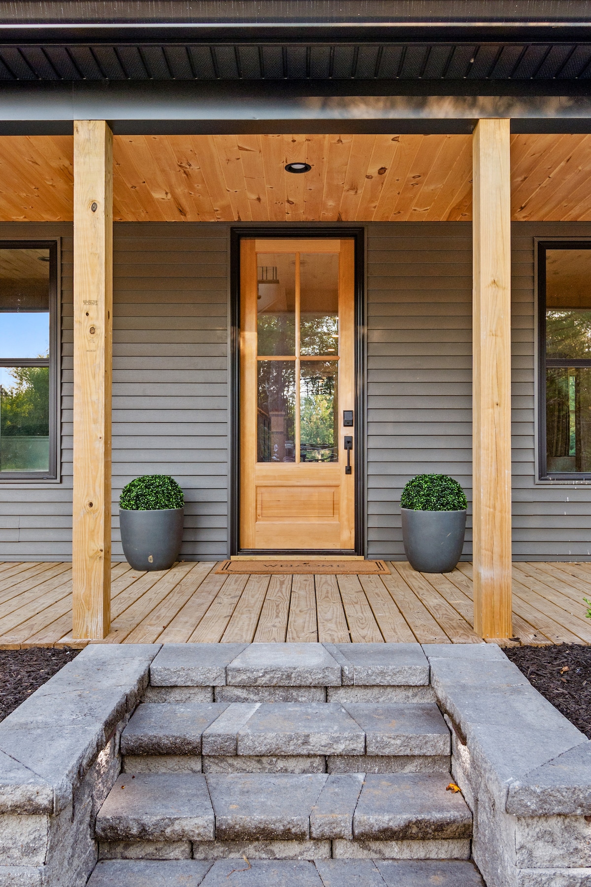 The entryway features a solid wood front door with vertical paneling, flanked by two potted shrubs. A spacious wooden deck leads to the door, with stone steps providing access to the entry. Large windows on either side enhance natural light in the area.