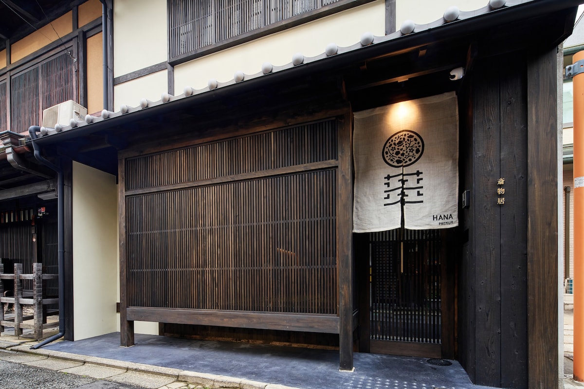 The entrance showcases traditional wooden architecture with horizontal slats and a distinct fabric banner displaying 'HANA.' The pathway is lined with smooth stones, leading to a welcoming door within a serene and historical setting.