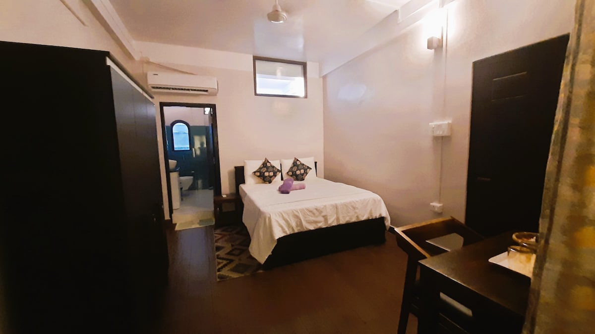 A well-lit room features a spacious bed with decorative pillows, complemented by a wooden desk and chair. A door leads to a bathroom, while natural light enters through a window, enhancing the minimalist decor and neutral-toned walls.