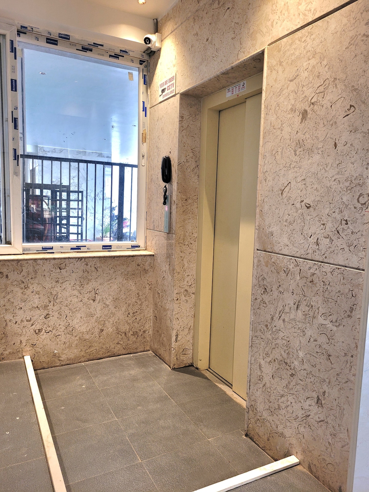 An elevator entrance is displayed with a beige textured panel wall, complemented by a gray tiled floor. Natural light is filtered through a large window nearby, showcasing a glimpse of the building's interior. Essential elevator controls are visible next to the door.