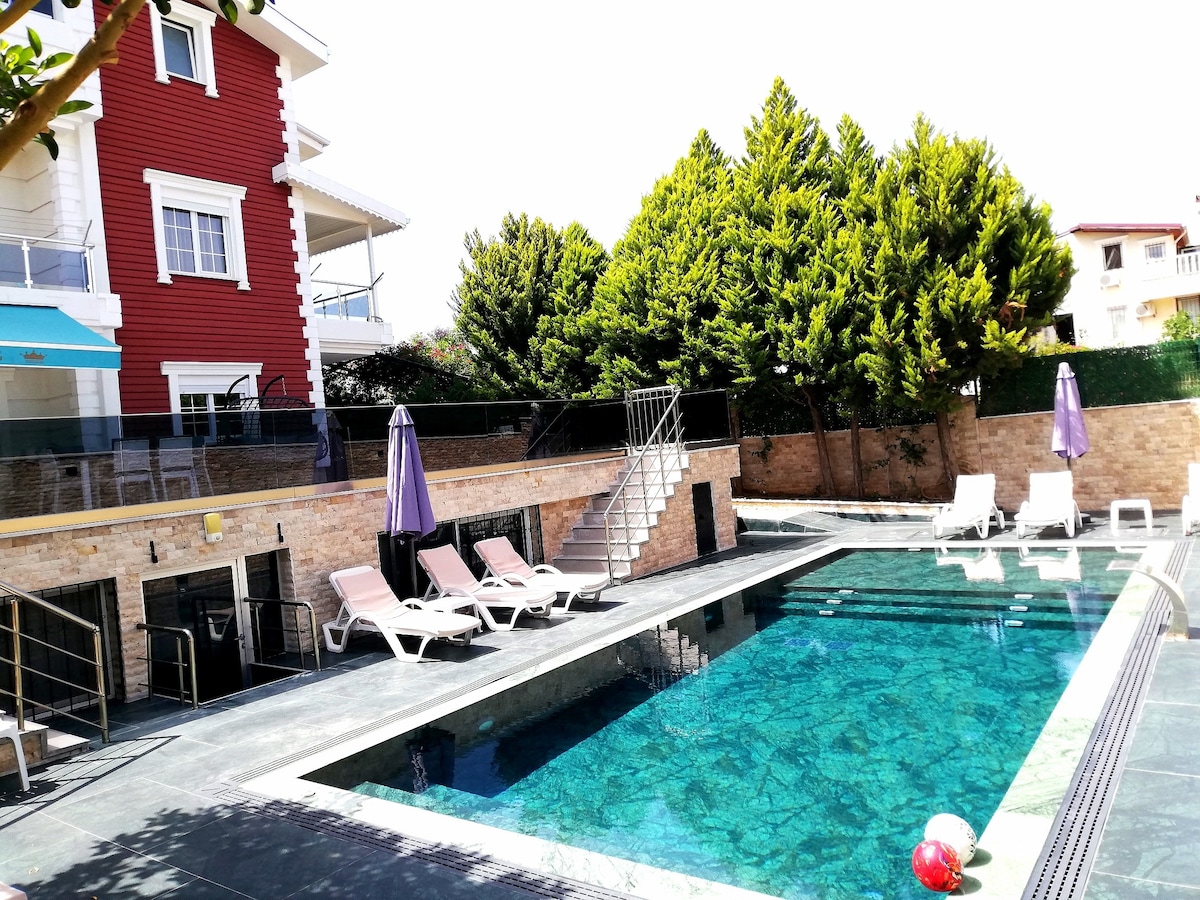 A spacious outdoor area features a heated pool surrounded by a sun terrace with lounge chairs. Vibrant greenery lines the backdrop, adding privacy. Two umbrellas provide shade, while steps lead to the villa entrance, complementing the elegant stonework of the surrounding walls.