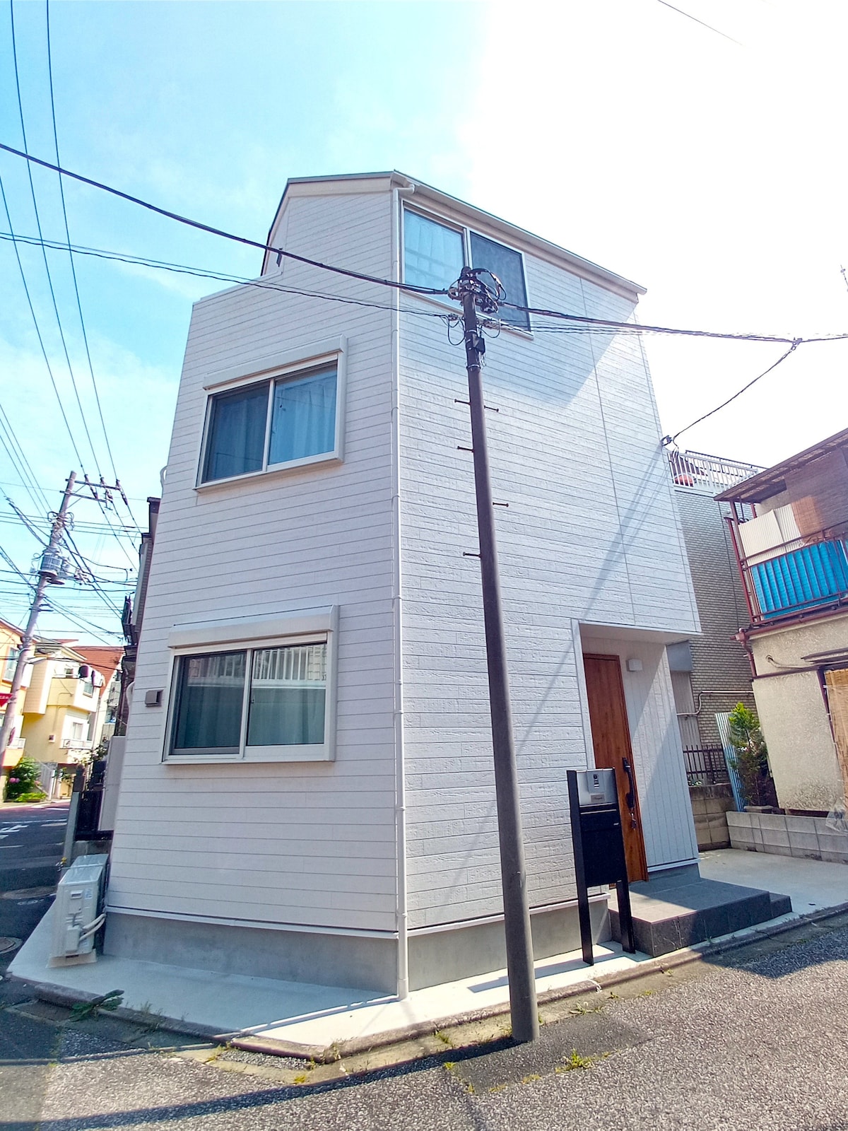 A modern, two-story house is situated in a quiet neighborhood. The exterior features a clean white facade and large windows allowing natural light. Nearby utility poles and power lines contribute to the urban landscape.