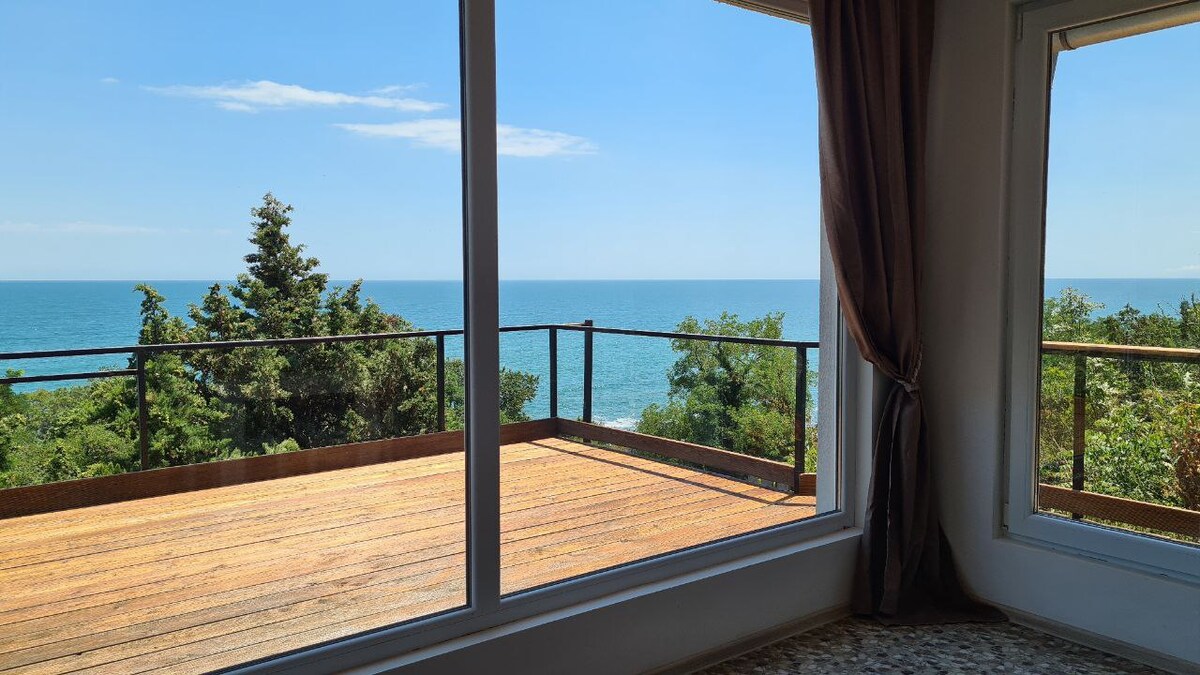 Large windows frame a beautiful view of the sea, inviting natural light into the room. A spacious terrace is visible outside, featuring a wooden deck that extends towards the horizon, surrounded by greenery. Soft curtains gently drape beside the windows, enhancing the serene atmosphere.