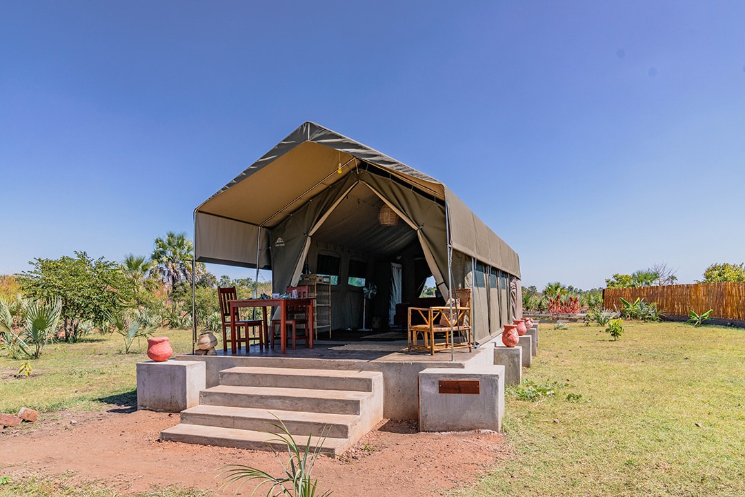 A spacious safari tent is situated on a raised platform, featuring a front porch with steps leading up to it. Outdoor dining furnishings are visible under the awning, surrounded by a lush garden and distant greenery. The tent is designed for comfort and connection with nature.