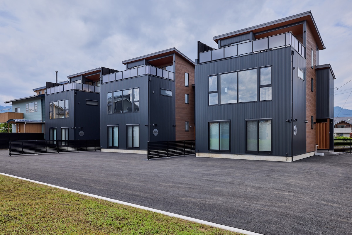 The exterior of a newly built three-story private villa is featured, showcasing a modern design with a combination of dark and wooden panels. Large windows allow natural light to fill the spaces. A spacious parking area is located in front, surrounded by a well-maintained grassy area.