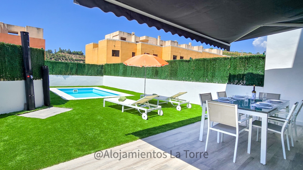 An outdoor space features an artificial grassy area with a private pool and sun loungers. A shaded dining table is arranged nearby, with a large orange umbrella providing protection from the sun. The enclosure is surrounded by tall green hedges, ensuring privacy.