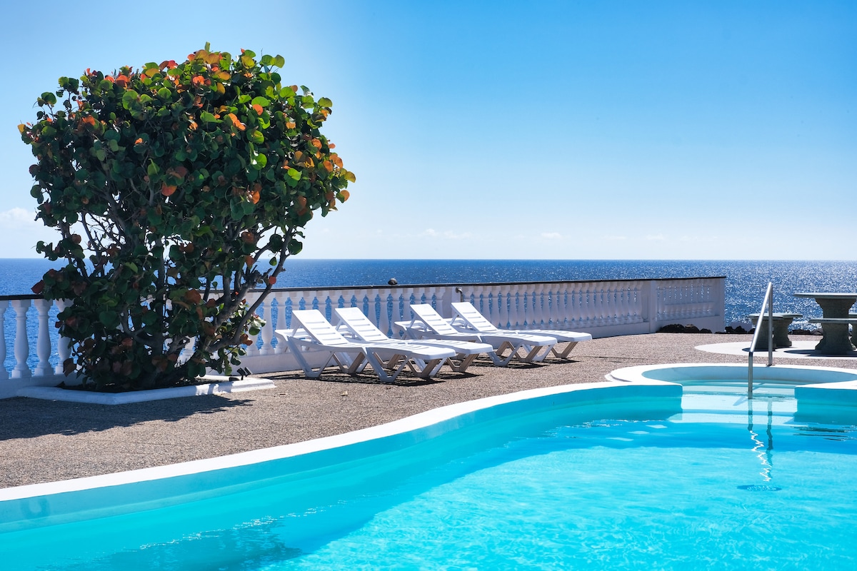 A solar-heated swimming pool is surrounded by a patio area and white lounge chairs. A lush tree adds greenery near the water's edge, with the expansive expanse of the Atlantic Ocean visible in the background under a clear blue sky.