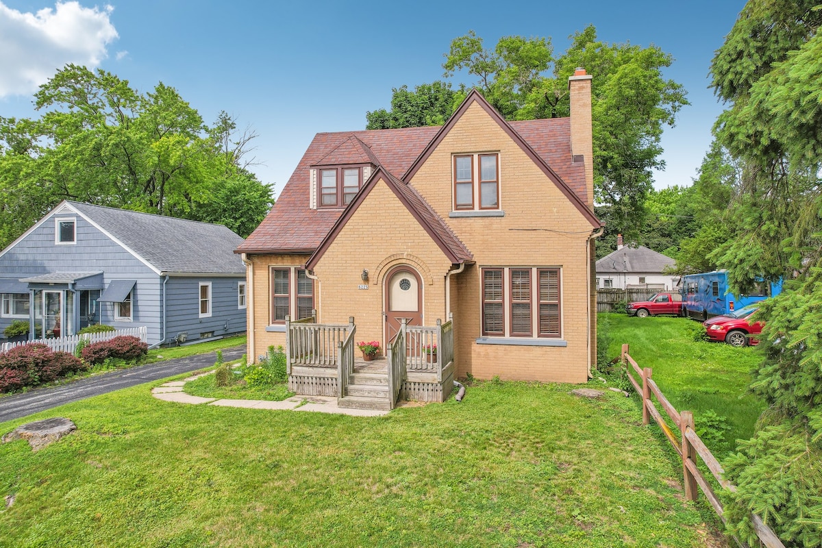 The charming vintage tudor home features a warm brick facade with a steeply pitched roof. A welcoming porch is complemented by wooden steps and railings, surrounded by a verdant lawn. Neighboring greenery and trees create a serene setting, enhancing the exterior appearance.
