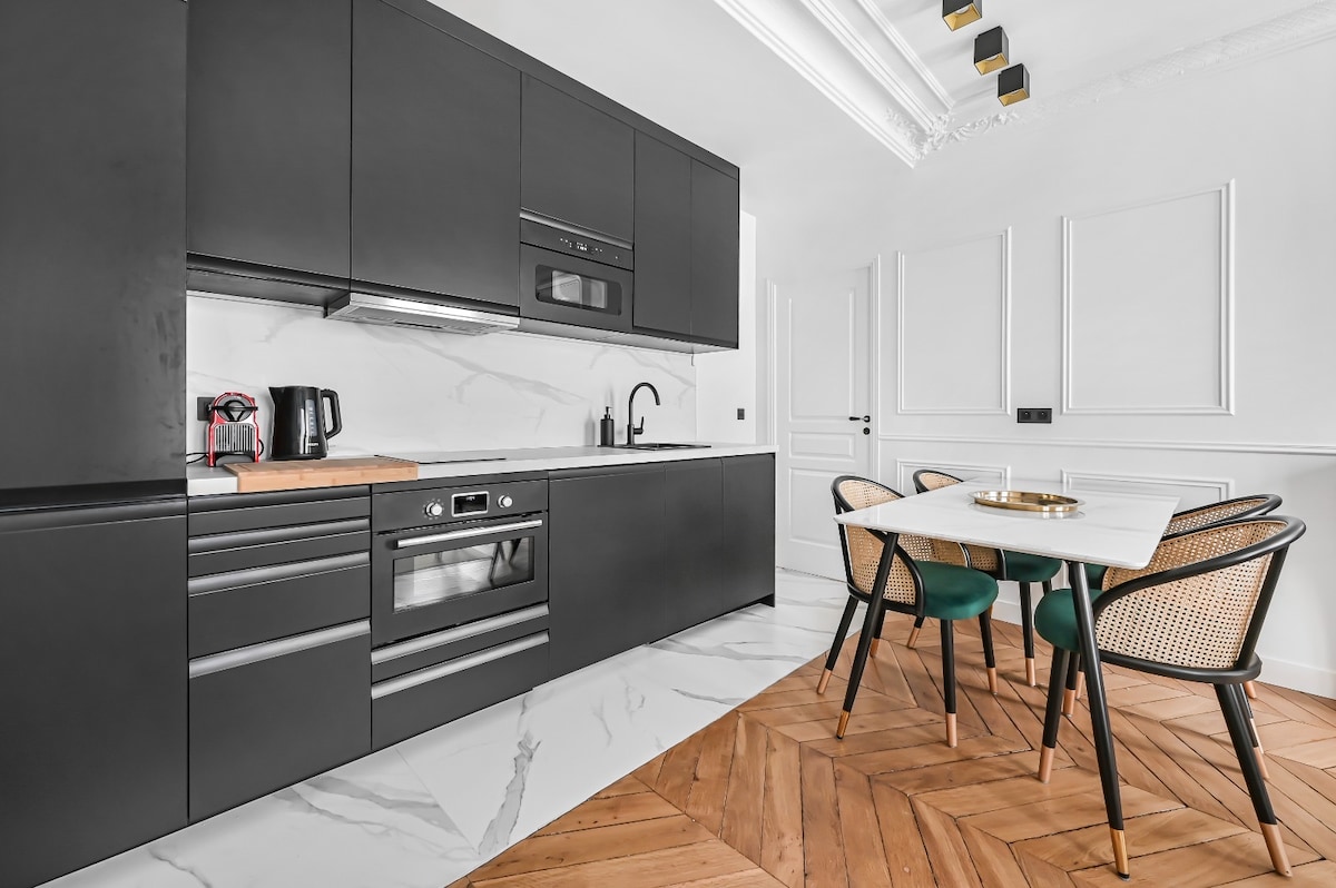 A modern kitchen features sleek black cabinetry and marble countertops. Built-in appliances include an oven and microwave. A wooden cutting board is placed on the counter, alongside a coffee maker. A dining table with four black and green chairs is positioned on a herringbone wooden floor.