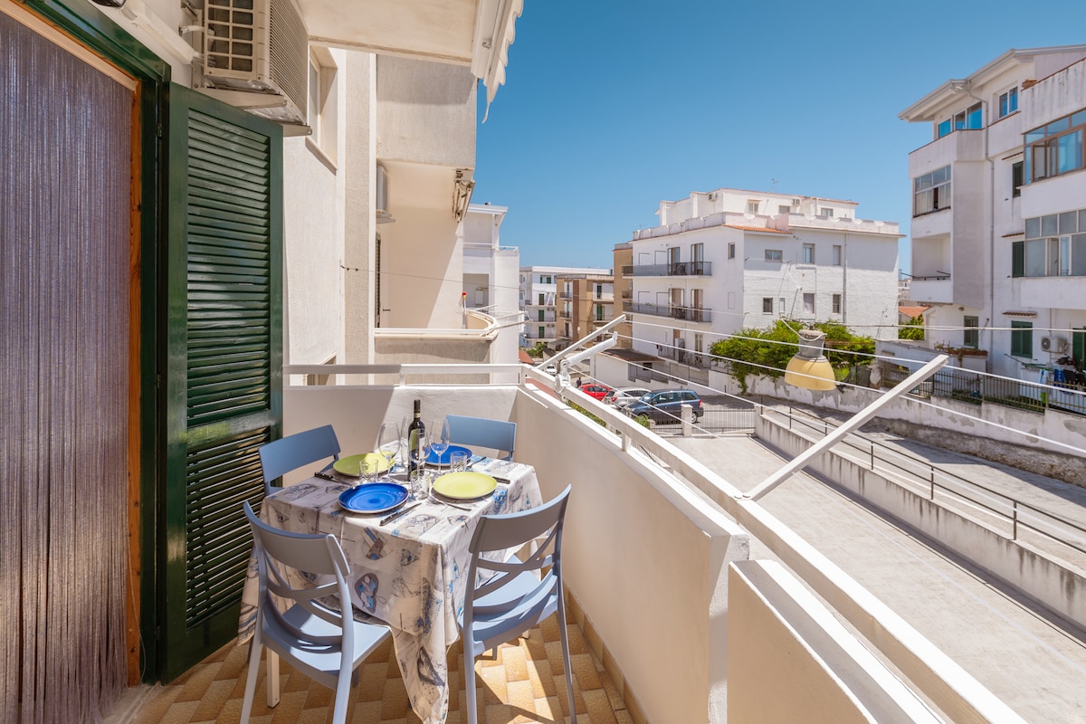 A balcony is shown featuring a round table set with blue plates and glasses. The surrounding area includes white buildings and a view of the street below, with clear blue skies overhead.
