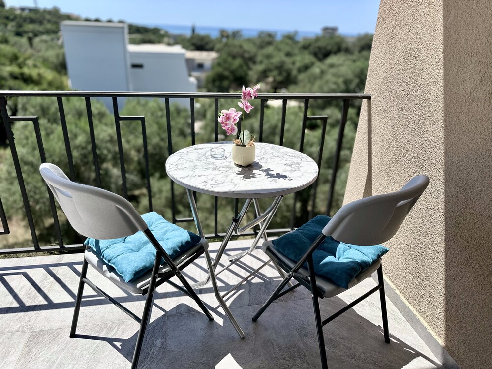 A small balcony features a round marble table paired with two foldable chairs, adorned with teal cushions. A potted plant with pink flowers adds a touch of color, while a scenic view of trees and the distant sea is visible in the background.