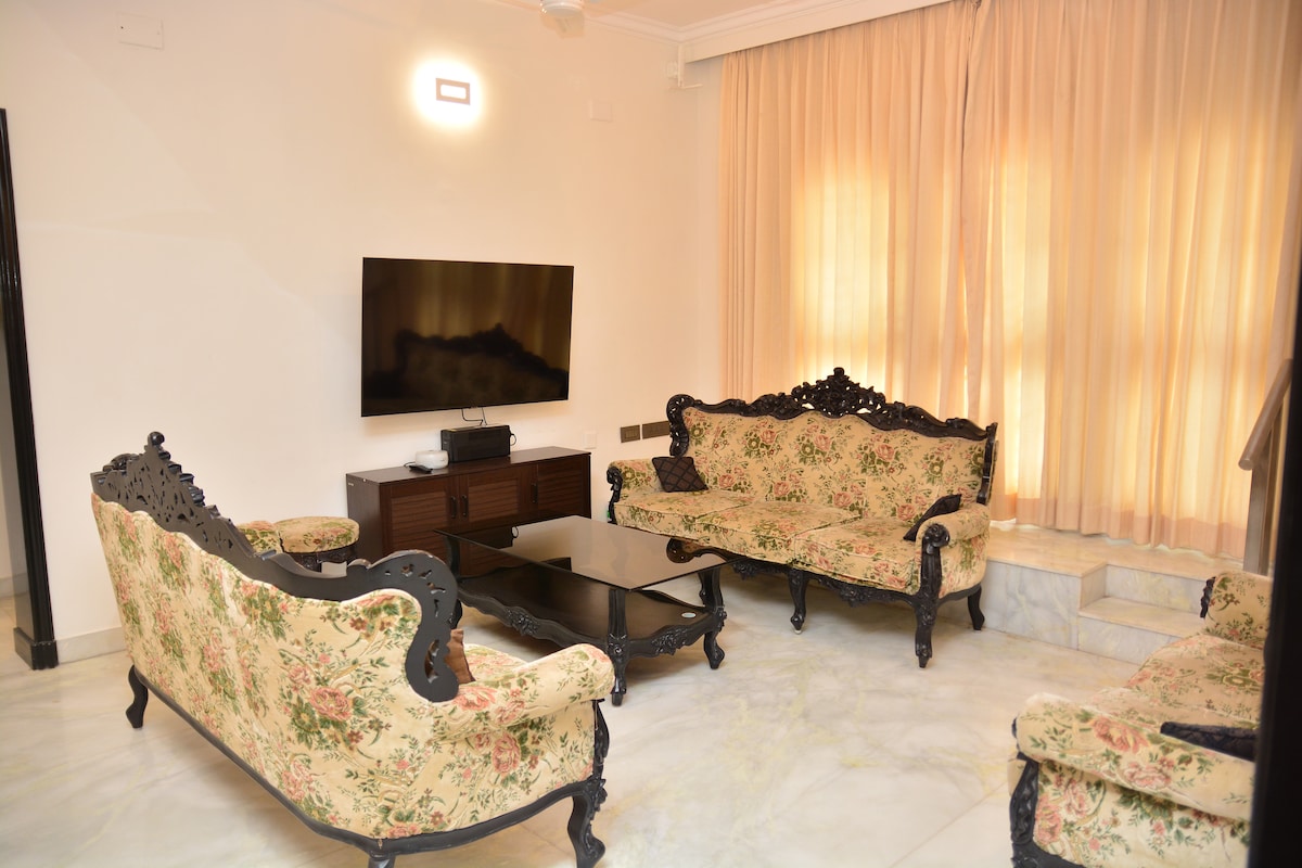 A spacious living area features elegant floral-patterned seating arrangements, including a pair of carved wooden sofas and a central coffee table. A wall-mounted television is positioned above a minimalist console. Soft, natural light filters through sheer curtains, complementing the neutral color palette.