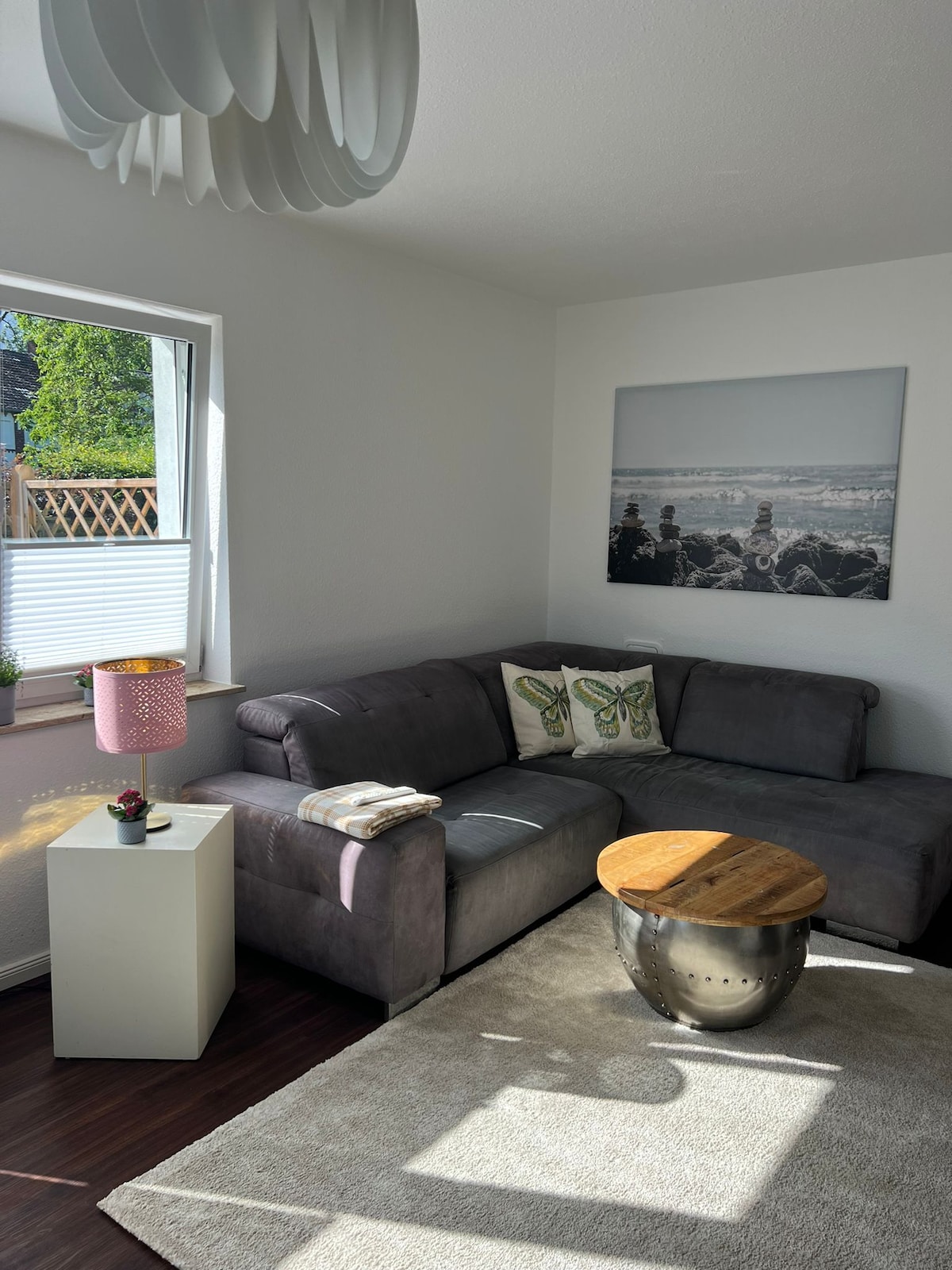 A comfortable living area is featured with a gray sofa adorned with decorative pillows. Natural light enters through the window, highlighting a small wooden side table and a light-colored rug beneath. A black-and-white artwork of a seaside scene is displayed on the wall.