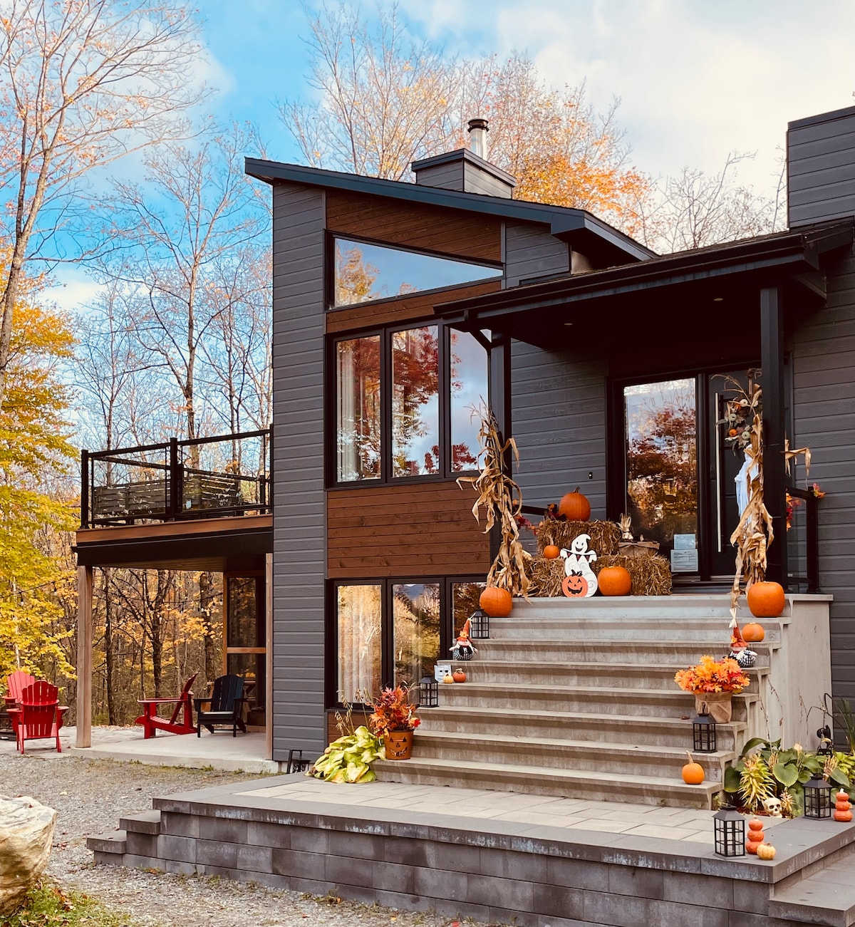 The exterior of the chalet features a contemporary design with large windows allowing ample natural light. A set of stairs, adorned with seasonal decorations including pumpkins and plants, leads to the main entrance. A cozy seating area with bright red chairs is visible to the side.