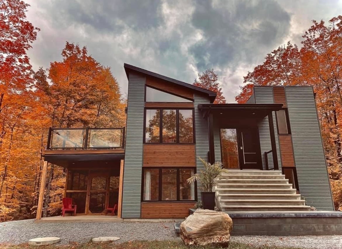 The modern chalet features an asymmetrical design, showcasing large windows that bring in natural light. A welcoming entrance with a staircase leads to a covered deck, surrounded by autumn foliage. The combination of wood and metal elements adds an inviting touch to the exterior.