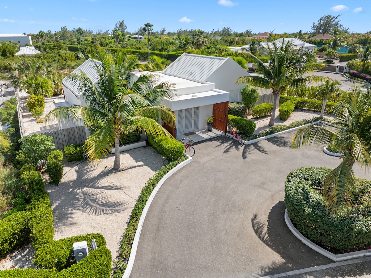 The exterior of Beach Landing Villa is shown, with tropical palm trees framing the entrance. A bicycle is positioned by the pathway leading to the front door. Lush greenery and neatly arranged landscaping contribute to the welcoming appearance of the property.