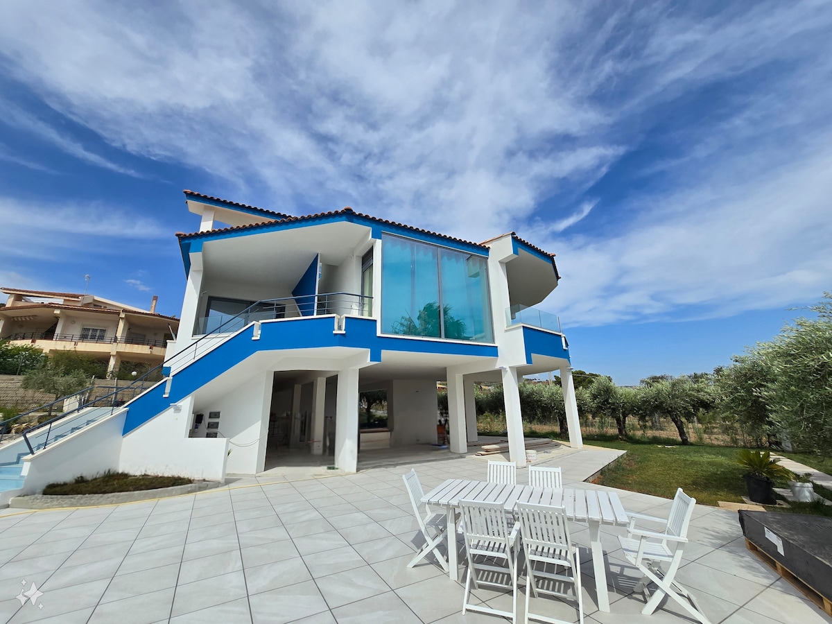 The exterior of the villa features a modern design with large glass windows and a striking blue accent. A spacious patio area is complemented by a white outdoor dining set, surrounded by lush greenery and a clear blue sky overhead.