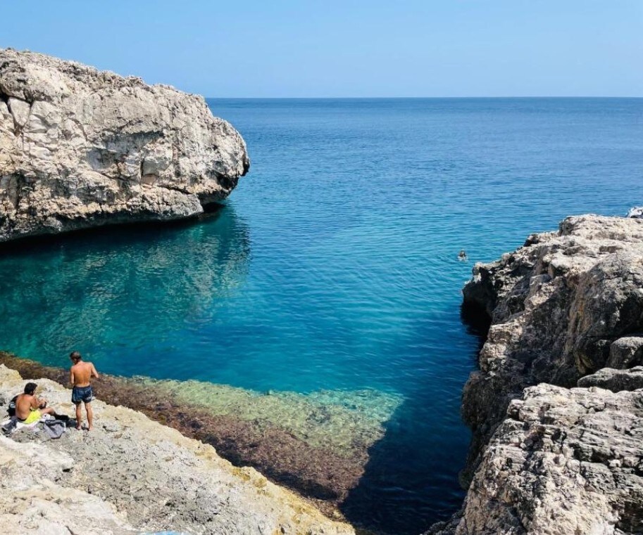 A rocky coastline is captured under clear blue skies, with calm turquoise waters lapping against the cliffs. Two individuals are seen near the water's edge, while a distant swimmer enjoys the serene surroundings. The rugged landscape provides a natural, peaceful retreat.