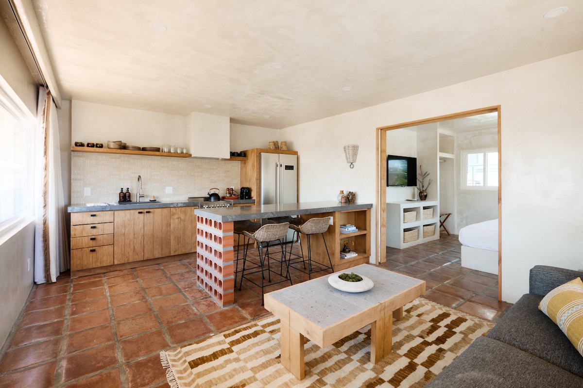 An open-concept living area features a modern kitchen with natural wood cabinetry and stainless steel appliances. A spacious dining counter with bar stools complements a rustic coffee table. Large windows provide ample natural light and a view of the surrounding area, with terracotta tiles adding warmth.