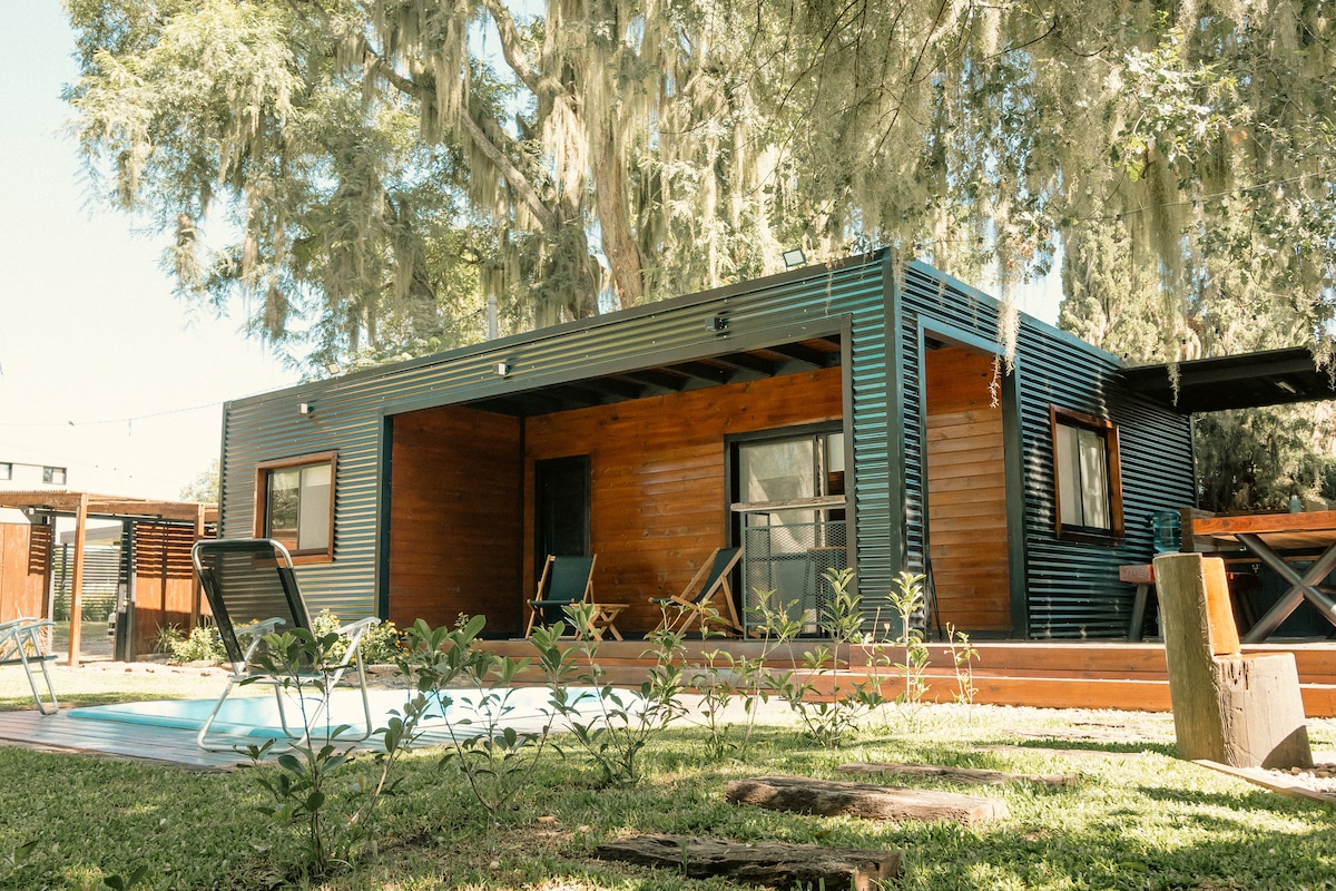 A modern cabin is set amidst lush greenery, featuring a wooden porch with two lounge chairs. Large windows allow natural light to fill the interior, while the surrounding landscaped area includes a swimming pool and well-maintained garden pathways.