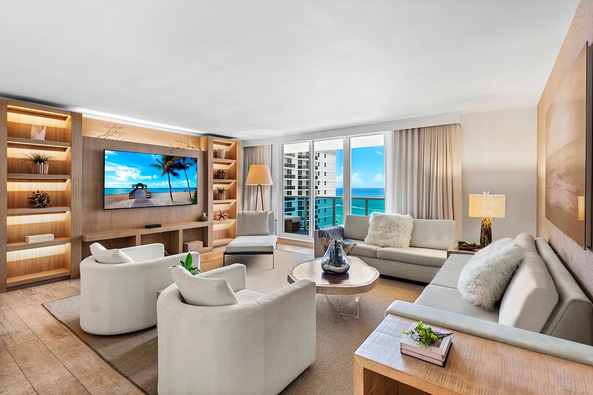 A bright living room showcases large glass doors that offer unobstructed ocean views. Two comfortable armchairs and a spacious sectional sofa are arranged around a decorative coffee table. A large flat-screen TV is positioned above a wooden media unit, which is complemented by softly glowing shelving.