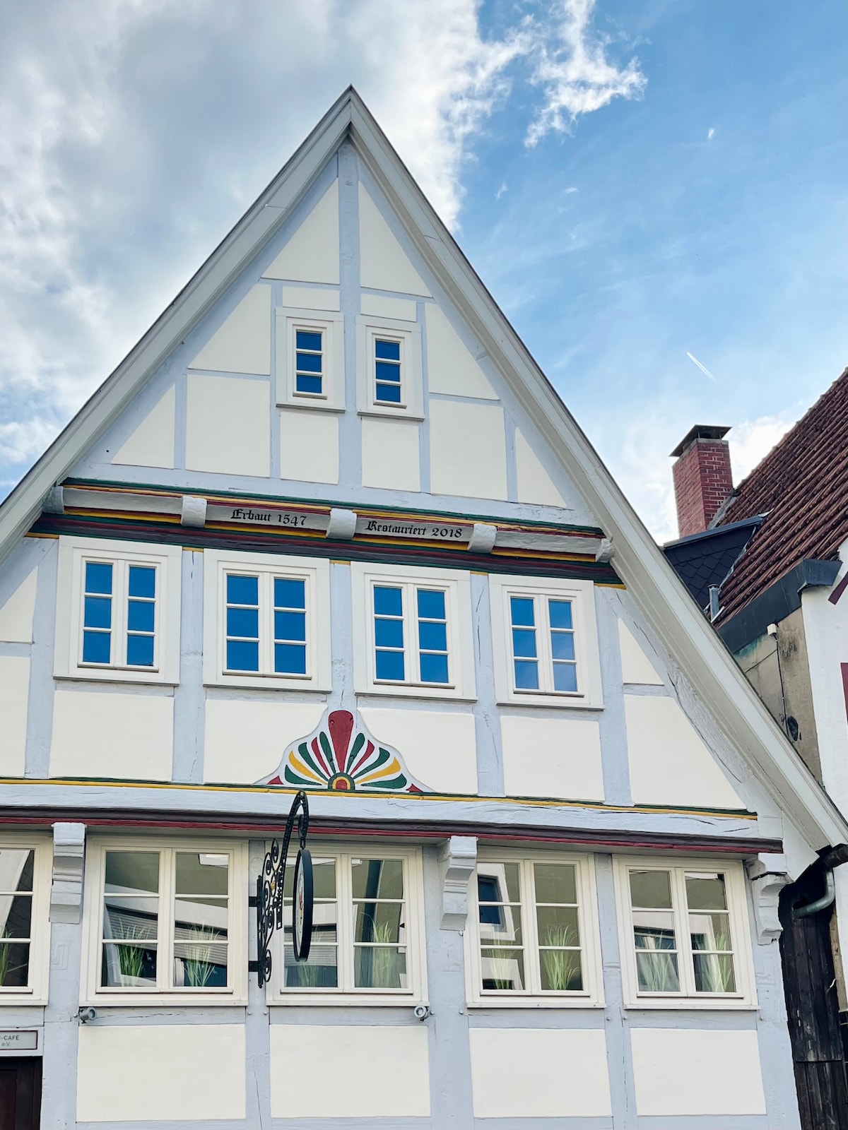 The exterior of a traditional half-timbered house is displayed, characterized by its distinctive triangular roof and white façade. Colorful decorative features are visible under the roofline, alongside multiple windows arranged symmetrically. A clear blue sky provides a bright backdrop to the charming architecture.