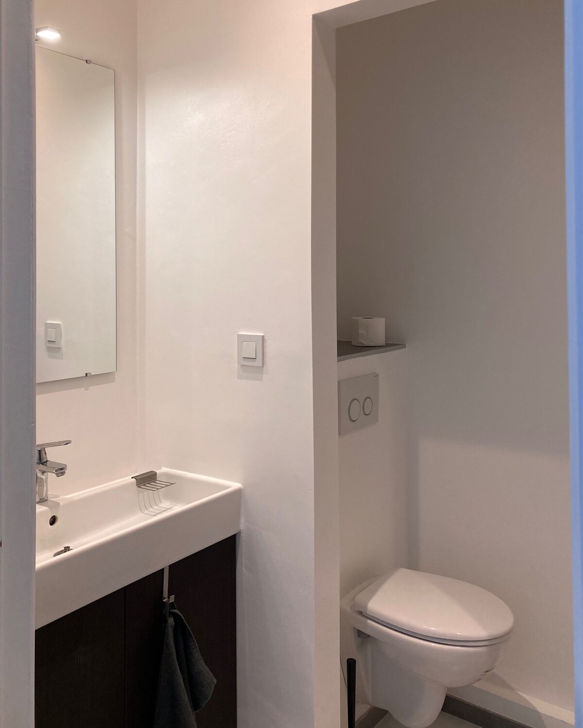 A compact bathroom area is presented, featuring a minimalist sink with a mirror above it. A toilet is positioned beside a small shelf, which includes a few toiletries. The walls are painted white, providing a clean and bright appearance.