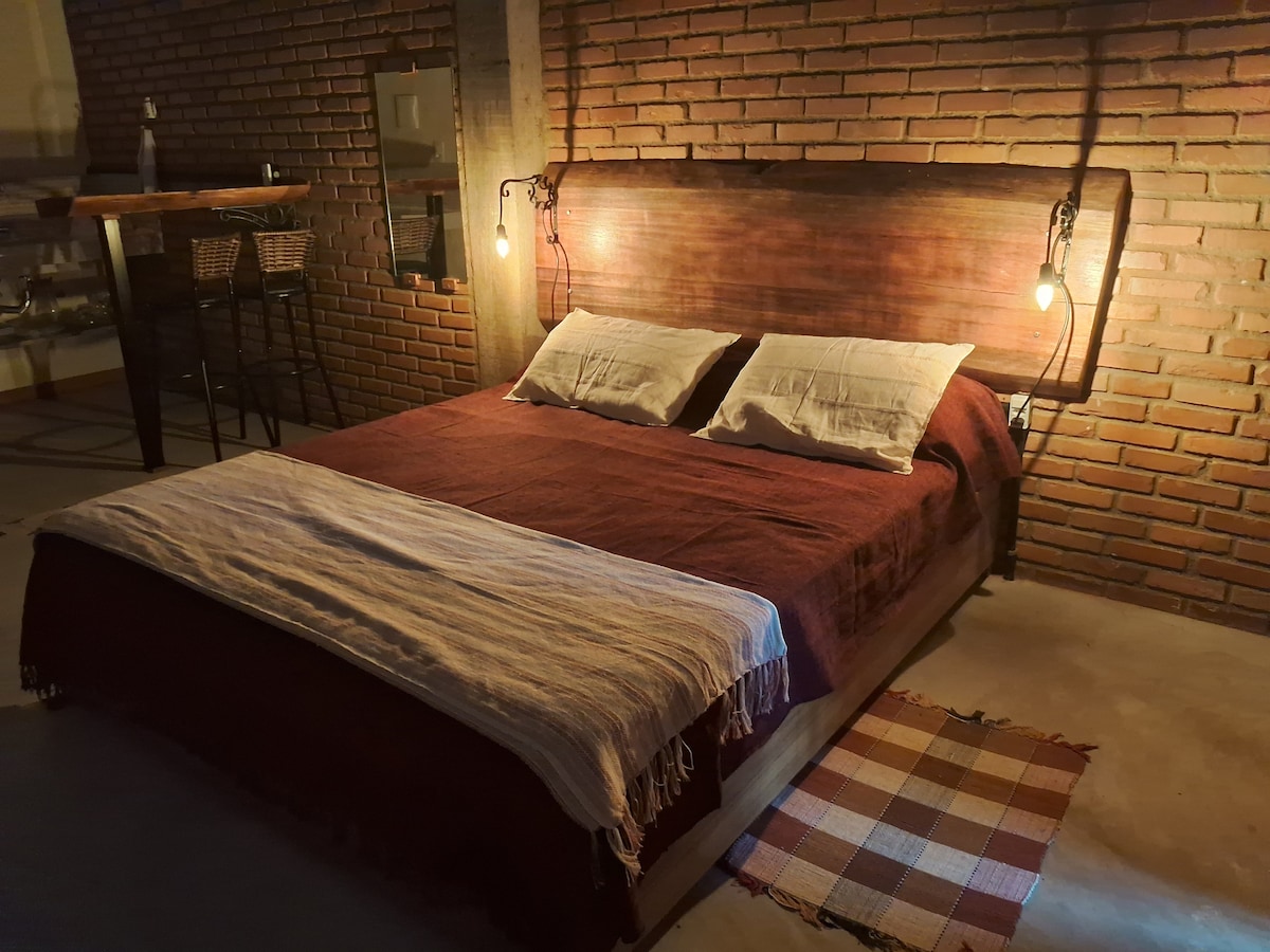 A spacious bed is positioned against a brick wall, featuring a wooden headboard and soft bedding. Two pendant lights illuminate the area, highlighting a woven throw at the foot of the bed. A small patterned rug is placed beside the bed, adding texture to the floor.