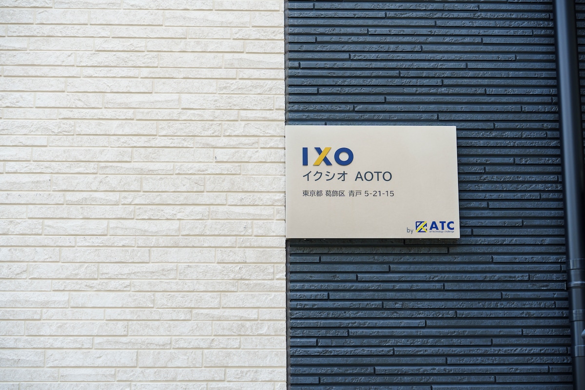 The image displays the exterior sign of the building, showing the name 'IXO AOTO.' The sign features a clean design with contrasting colors against the brick backdrop of the building, which alternates between dark and light tones.
