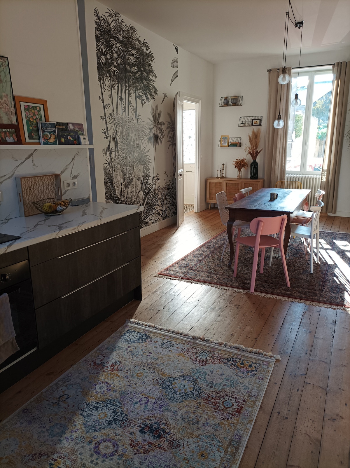 A dining area features a long wooden table surrounded by several chairs in varying colors. A large rug underlines the space, adding texture. Natural light enters through a tall window, highlighting a wall adorned with a botanical mural and decorative artwork.