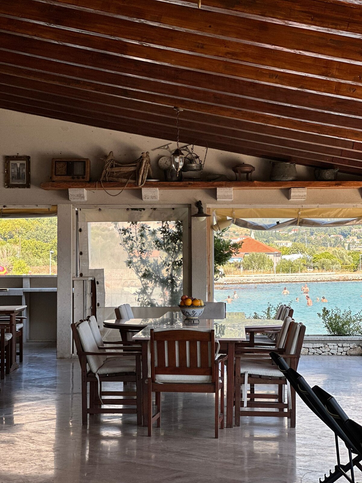 The spacious dining area features a wooden table surrounded by six chairs, situated beneath a beam ceiling. Natural light floods the space, and views of the nearby water can be seen through large windows, enhancing the inviting atmosphere.
