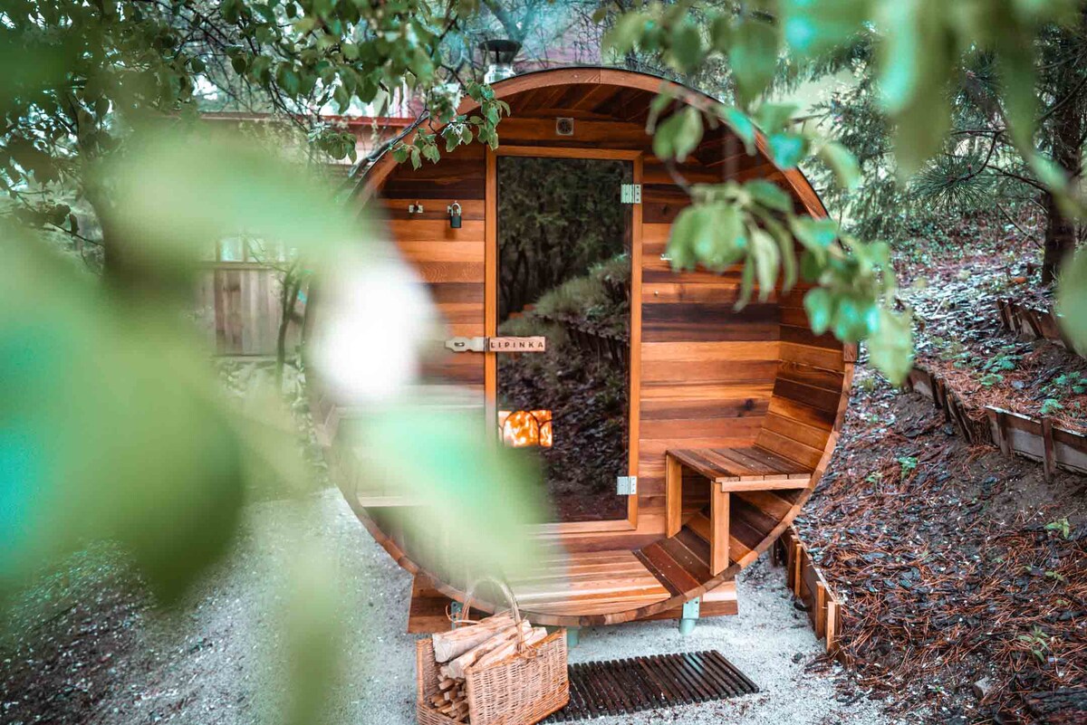 A wooden sauna, shaped like a barrel, is situated in a natural setting surrounded by greenery. The entrance features a clear glass door and wooden benches, while a woven basket rests at the base, inviting relaxation and wellness.