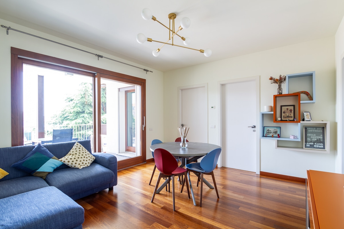 A bright living area showcases a comfortable sofa adorned with colorful cushions. A round dining table is surrounded by four chairs. Large glass doors offer access to the terrace, while wooden flooring adds warmth to the space.