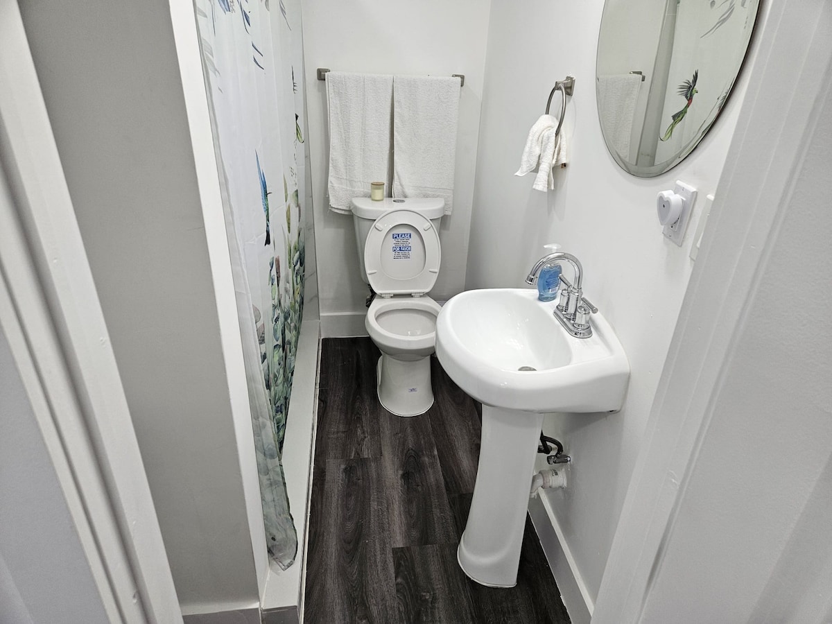 A modern bathroom is shown, featuring a clean toilet, a sink with a sleek faucet, and a round mirror above it. A colorful shower curtain provides privacy while white towels hang neatly on a rack. The flooring is dark, complementing the light-colored walls.