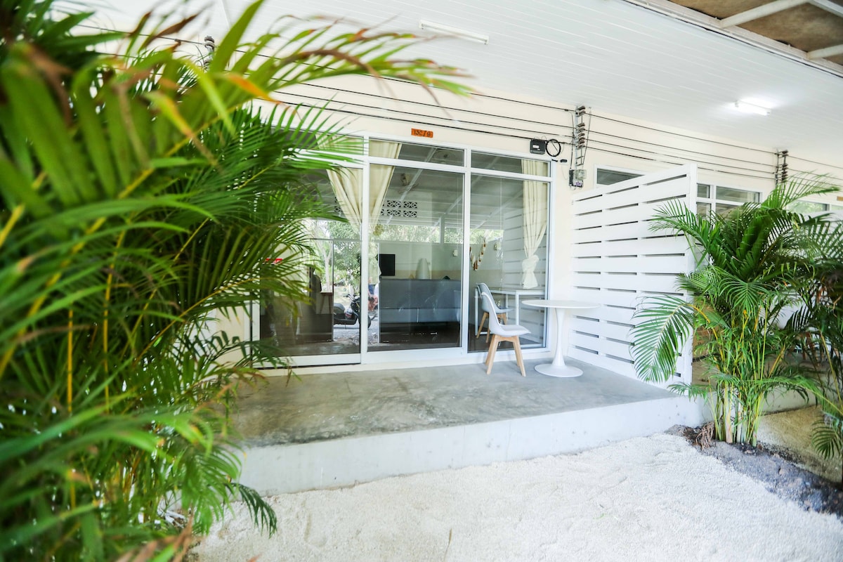 An entrance area reveals a modern apartment with large glass doors, framed by lush green plants. A small table and chair sit on the patio, set against a sandy ground that adds a relaxing touch to the outdoor space.